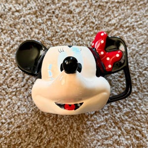 May include: A ceramic mug shaped like Minnie Mouse's face. The mug features black ears, a red bow with white polka dots, and a black handle. The face is white with black eyes, a black nose, and a red mouth.