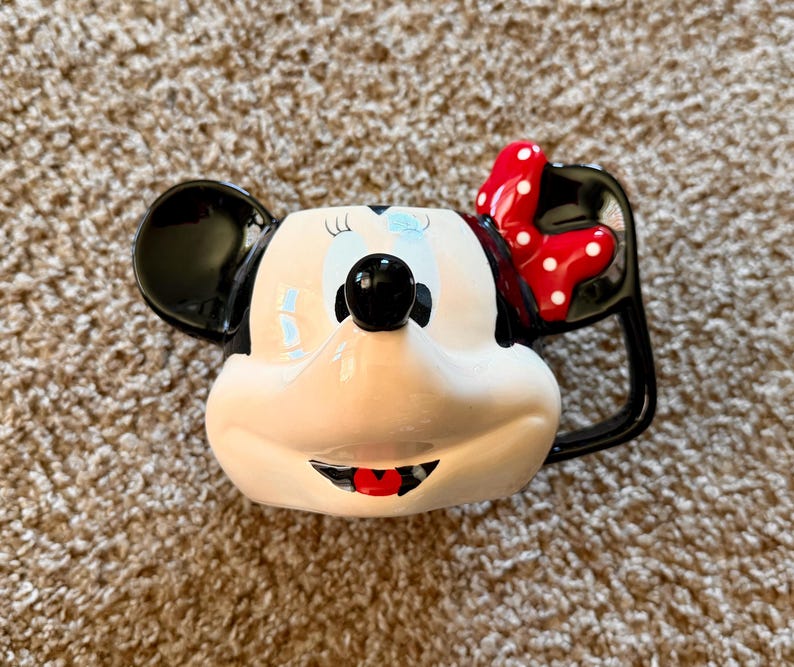 May include: A ceramic mug shaped like Minnie Mouse's face. The mug features black ears, a red bow with white polka dots, and a black handle. The face is white with black eyes, a black nose, and a red mouth.