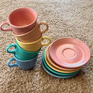 May include: A stack of four ceramic teacups and a stack of four matching saucers in pastel colours including pink, peach, yellow, teal, and light blue. Each cup features a rounded handle and horizontal ribbed detailing along the exterior surface.