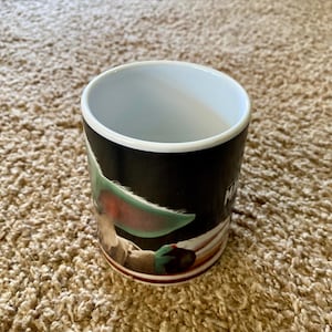 May include: A ceramic mug featuring a character from a popular science fiction series. The mug has a black background with a white rim and a brown base. The character has large green ears and brown eyes. The word "M" is visible on the side of the mug.