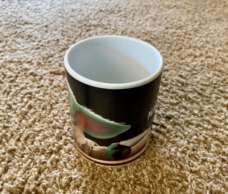 May include: A ceramic mug featuring a character from a popular science fiction series. The mug has a black background with a white rim and a brown base. The character has large green ears and brown eyes. The word "M" is visible on the side of the mug.