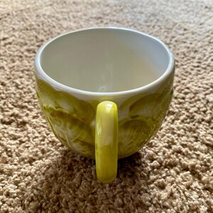 May include: A ceramic mug with a light green exterior and a yellow handle. The mug's exterior features a textured design, and the interior is white. The mug is designed for hot beverages.
