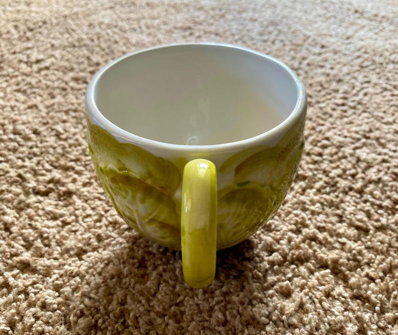 May include: A ceramic mug with a light green exterior and a yellow handle. The mug's exterior features a textured design, and the interior is white. The mug is designed for hot beverages.