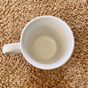 Puede incluir: Vista a&eacute;rea de una taza de cer&aacute;mica blanca con un asa curva. El interior de la taza es de color crema claro. La taza est&aacute; sobre una superficie marr&oacute;n texturizada.