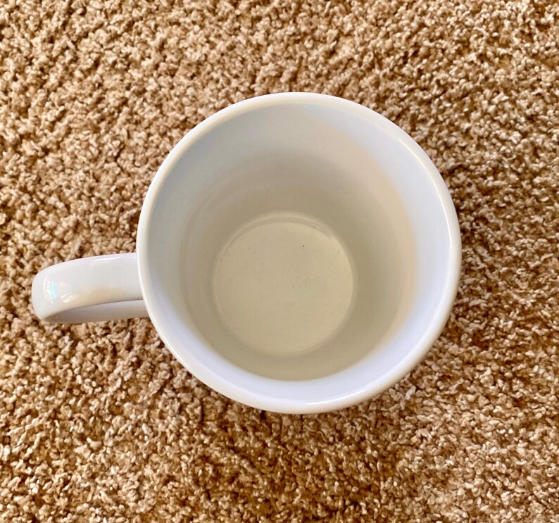 Puede incluir: Vista a&eacute;rea de una taza de cer&aacute;mica blanca con un asa curva. El interior de la taza es de color crema claro. La taza est&aacute; sobre una superficie marr&oacute;n texturizada.