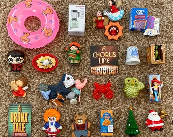 May include: A collection of miniature toys and figurines, including a pink inflatable ring with duck designs, a miniature blender box, a Harry Potter figurine, a miniature musical theater poster, and a Christmas tree.
