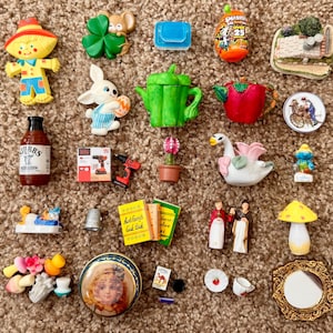 May include: A collection of miniature figurines and trinkets, including a scarecrow, a bunny, a teapot, a bottle of sauce, a drill, a swan, a mushroom, and a small book. The items are arranged on a brown, textured surface.