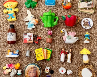 May include: A collection of miniature figurines and trinkets, including a scarecrow, a bunny, a teapot, a bottle of sauce, a drill, a swan, a mushroom, and a small book. The items are arranged on a brown, textured surface.