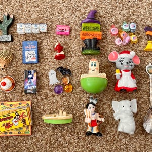 Puede incluir: Una colección de figuritas y baratijas en miniatura, que incluyen un cactus, un erizo, una bruja, un ratón con gorro de Papá Noel y una figura nativa americana. Otros artículos incluyen un diario en miniatura, una cinta de cassette y un juego.