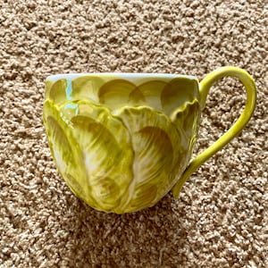 May include: A ceramic mug with a yellow handle and a textured, layered design in shades of yellow and white. The mug has a unique, organic shape and is set against a neutral background.
