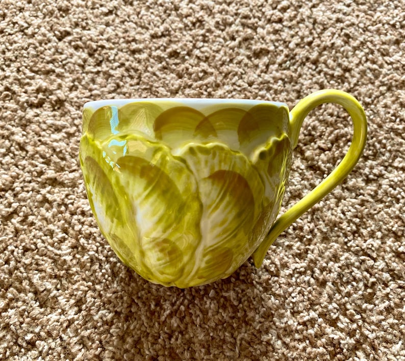 May include: A ceramic mug with a yellow handle and a textured, layered design in shades of yellow and white. The mug has a unique, organic shape and is set against a neutral background.