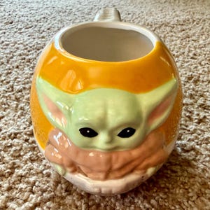 May include: A ceramic mug with a rounded shape and a handle. The mug features a light green and tan character design on an orange background. The character has large ears and black eyes.