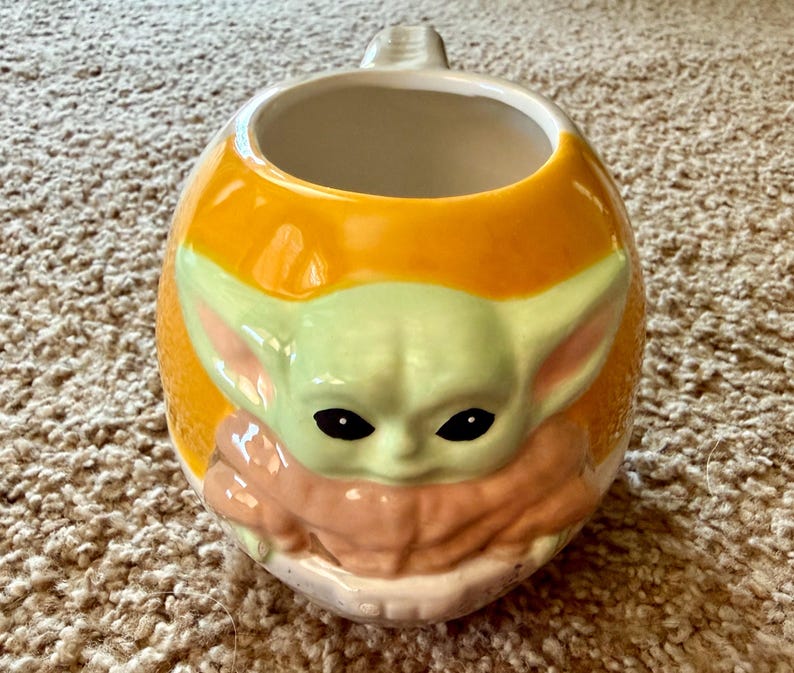 May include: A ceramic mug with a rounded shape and a handle. The mug features a light green and tan character design on an orange background. The character has large ears and black eyes.