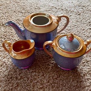 以下が含まれることがあります： ティーポット、クリーマー、砂糖入れの3点セット。青と金のイリデセント仕上げです。ティーポットには注ぎ口と取っ手があり、砂糖入れには蓋と取っ手があります。クリーマーには取っ手があります。