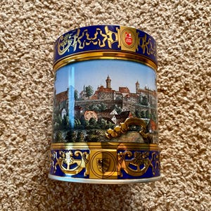 May include: A blue and gold tin featuring a picture of a town. The tin has a gold trim and a gold crest on the front. The town is surrounded by trees and a river.