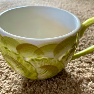 May include: A ceramic coffee mug with a white interior and a lime green handle. The exterior features a hand-painted design of overlapping green brushstrokes. The mug is designed for hot beverages.