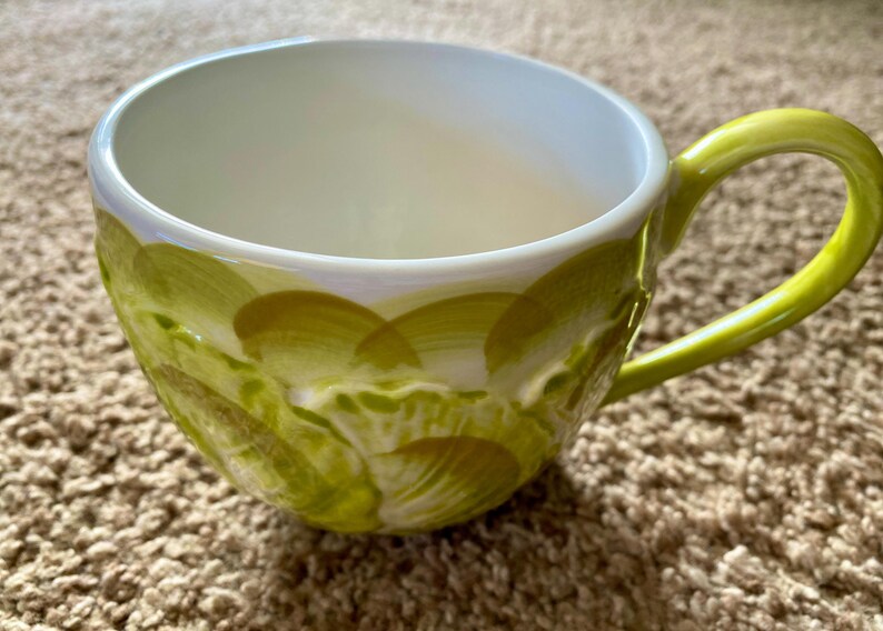 May include: A ceramic coffee mug with a white interior and a lime green handle. The exterior features a hand-painted design of overlapping green brushstrokes. The mug is designed for hot beverages.