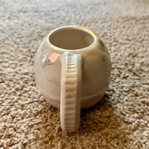 May include: Cream-colored ceramic mug with a rounded body and a textured handle. The mug has a wide opening and a slightly distressed finish with dark speckles. The handle is a solid, rectangular shape with vertical ridges.