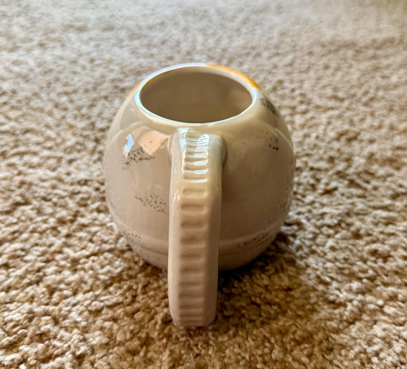 May include: Cream-colored ceramic mug with a rounded body and a textured handle. The mug has a wide opening and a slightly distressed finish with dark speckles. The handle is a solid, rectangular shape with vertical ridges.