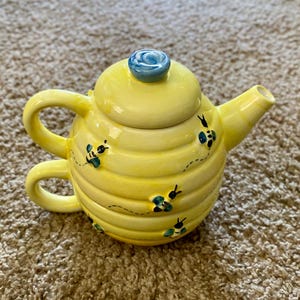 Puede incluir: Una tetera de cerámica amarilla con tapa y asa azules. La tetera está decorada con ilustraciones de abejas negras y azules y un diseño de panal. La tetera tiene un pico y un asa.
