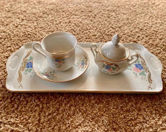 Może przedstawiać: Biały porcelanowy zestaw do herbaty z kwiatowymi wzorami. Zestaw zawiera filiżankę do herbaty, spodek i cukiernicę z pokrywką. Zestaw znajduje się na białej tacy z pozłacanymi zdobieniami.