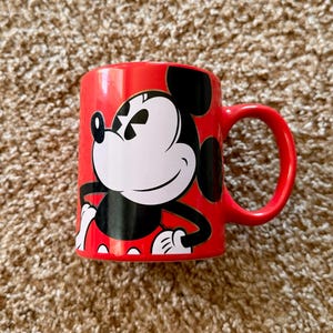 K&ouml;nnte beinhalten: Eine rote Keramik-Tasse mit einem Mickey Mouse-Design. Die Tasse hat einen abgerundeten Henkel und eine gl&auml;nzende Oberfl&auml;che. Die ikonische Figur ist in Schwarz und Wei&szlig; vor rotem Hintergrund dargestellt, ein klassisches Disney-Sammlerst&uuml;ck.