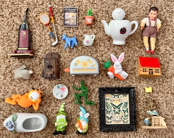 Puede incluir: Una colección de objetos decorativos en miniatura, que incluyen una aspiradora, una tetera, una muñeca, una tostadora, un marco de fotos y varias figuras. Los artículos están dispuestos sobre una superficie marrón texturizada.