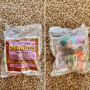May include: Two sealed McDonald's Happy Meal bags featuring Peanuts characters. One bag displays the text "PEANUTS" and "LINUS MILK MOVER". The other bag contains a Peanuts toy. The bags are on a brown carpet.