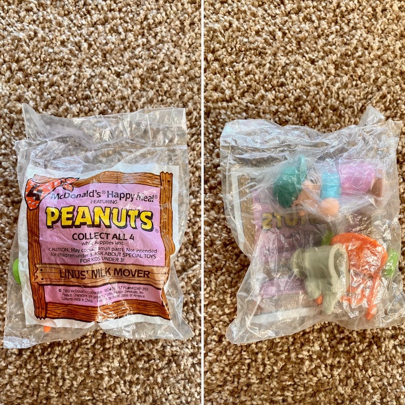 May include: Two sealed McDonald's Happy Meal bags featuring Peanuts characters. One bag displays the text "PEANUTS" and "LINUS MILK MOVER". The other bag contains a Peanuts toy. The bags are on a brown carpet.