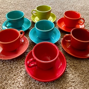 Puede incluir: Un juego de diez tazas de té y platillos de cerámica de colores. Las tazas y los platillos están dispuestos en círculo sobre una alfombra marrón. Las tazas son de diferentes colores, incluyendo rojo, azul, verde y amarillo. Los platillos son del mismo color que las tazas.
