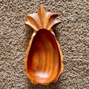 May include: A wooden pineapple-shaped bowl with a smooth, dark brown finish. The bowl is carved with a detailed pineapple design and has a deep, rounded interior.