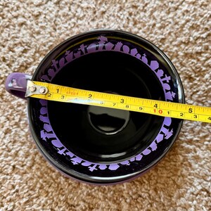 以下が含まれることがあります： 紫色の取っ手と縁が付いた黒いセラミックマグ。縁には紫色の墓石の繰り返し模様があります。マグカップの直径は約10cmです。