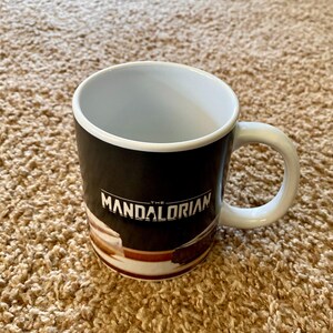 May include: A white-handled coffee mug with the text "THE MANDALORIAN" in white. The mug has a black background with a brown and white design at the bottom. The inside of the mug is white.