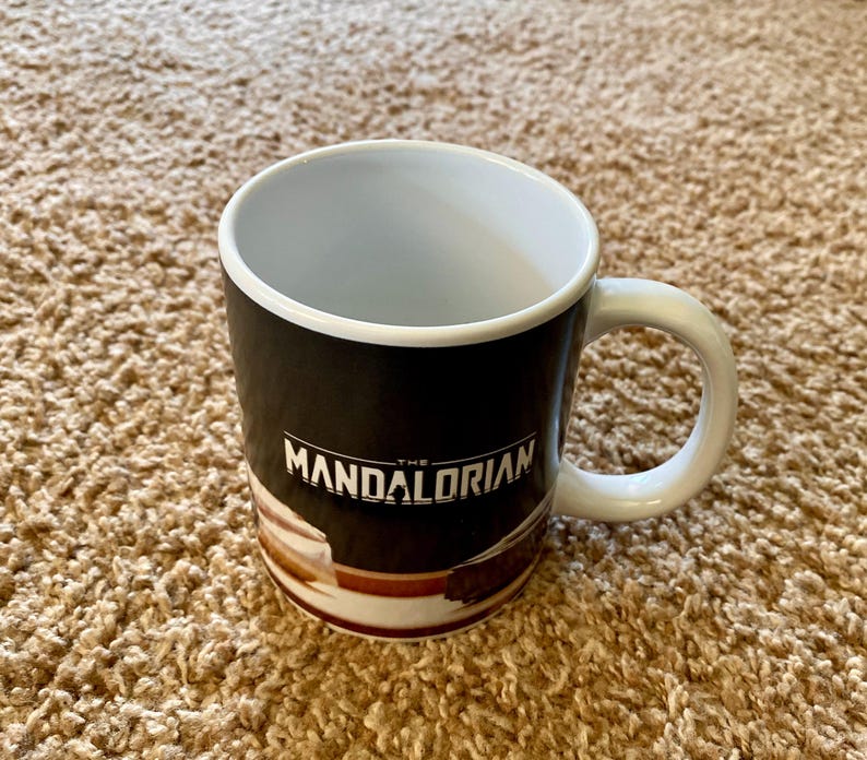 May include: A white-handled coffee mug with the text "THE MANDALORIAN" in white. The mug has a black background with a brown and white design at the bottom. The inside of the mug is white.