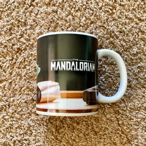May include: White ceramic mug with a black background featuring the text "THE MANDALORIAN" in white. The mug has a white handle and a brown and white design at the bottom. The mug is a Star Wars themed product.