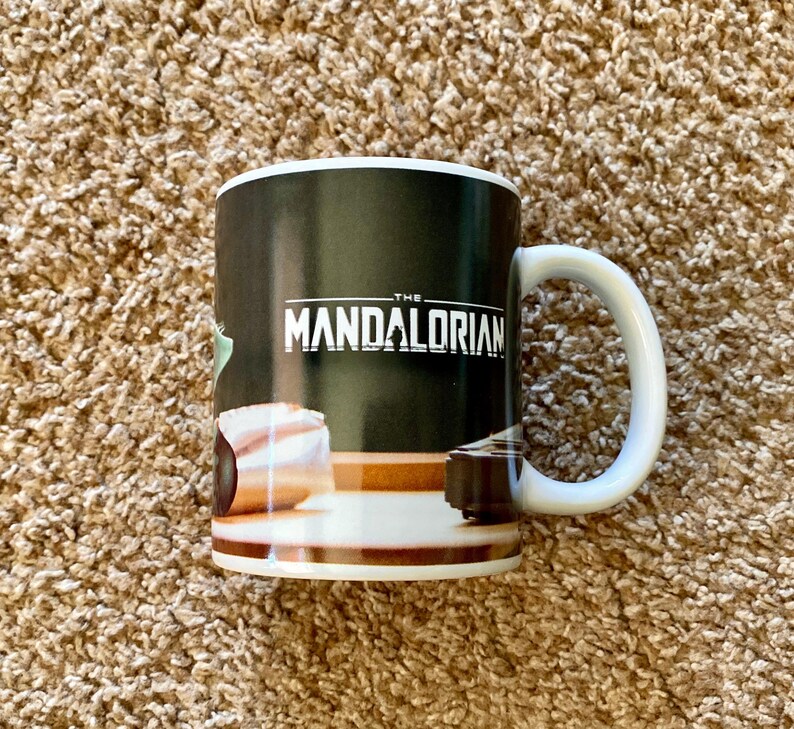 May include: White ceramic mug with a black background featuring the text "THE MANDALORIAN" in white. The mug has a white handle and a brown and white design at the bottom. The mug is a Star Wars themed product.