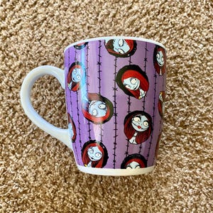 Puede incluir: Una taza de cer&aacute;mica con asa blanca y fondo morado con un patr&oacute;n repetido de Sally de Pesadilla antes de Navidad. El dise&ntilde;o incluye la cara de Sally y alambre de p&uacute;as negro.