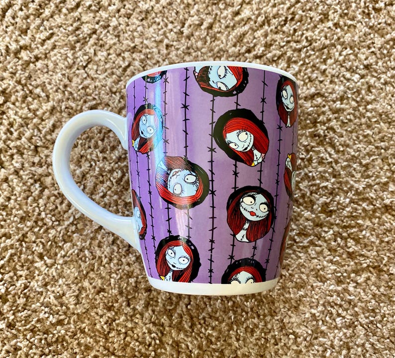 Puede incluir: Una taza de cer&aacute;mica con asa blanca y fondo morado con un patr&oacute;n repetido de Sally de Pesadilla antes de Navidad. El dise&ntilde;o incluye la cara de Sally y alambre de p&uacute;as negro.