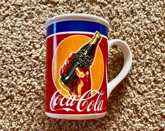 May include: White ceramic mug with a red, yellow, and blue Coca-Cola design. The design features a hand holding a bottle of Coca-Cola.