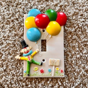 Puede incluir: Una cubierta decorativa para interruptor de luz con un diseño de payaso sosteniendo globos de colores. Los globos son rojos, amarillos, verdes y azules. El payaso lleva un sombrero de copa negro, un cuello con volantes azules y una chaqueta amarilla.