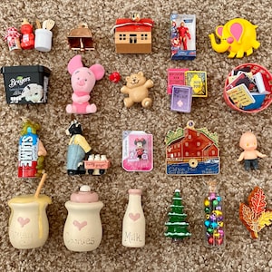 Puede incluir: Una colección de figuritas en miniatura y objetos decorativos, que incluyen una casa roja, un elefante amarillo, un cerdito rosa, un oso de peluche y un recipiente de helado en miniatura. Otros artículos incluyen una botella de leche, un árbol de Navidad y un tarro de caramelos de colores.