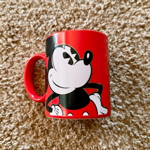 K&ouml;nnte beinhalten: Eine wei&szlig;e Keramiktasse mit schwarzem Henkel und dem Text "But First, Coffee" in fetter, schwarzer Schrift. Die Tasse steht vor einem verschwommenen Hintergrund, der eine gem&uuml;tliche Umgebung suggeriert.