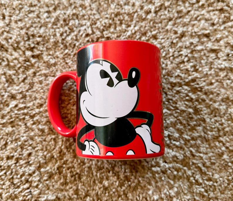 K&ouml;nnte beinhalten: Eine wei&szlig;e Keramiktasse mit schwarzem Henkel und dem Text "But First, Coffee" in fetter, schwarzer Schrift. Die Tasse steht vor einem verschwommenen Hintergrund, der eine gem&uuml;tliche Umgebung suggeriert.