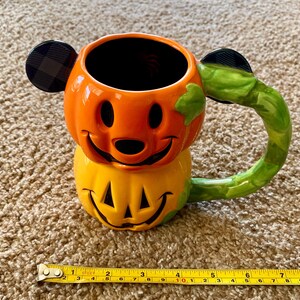 以下が含まれることがあります： ミッキーマウスの耳が付いたカボチャの形をしたセラミックハロウィーンマグ。マグはオレンジと黄色で、緑色のハンドルが付いています。耳は黒とチェック柄です。マグの高さは約10cmです。