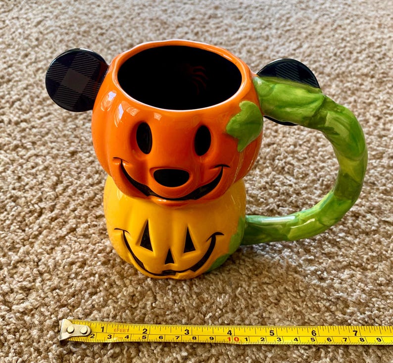 以下が含まれることがあります： ミッキーマウスの耳が付いたカボチャの形をしたセラミックハロウィーンマグ。マグはオレンジと黄色で、緑色のハンドルが付いています。耳は黒とチェック柄です。マグの高さは約10cmです。