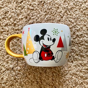 Könnte beinhalten: Weiße Keramiktasse mit Mickey Mouse in festlichem Urlaubsdesign. Mickey trägt einen rot-schwarz karierten Schal und rote Shorts. Die Tasse hat einen goldenen Griff und ist mit Weihnachtsbäumen und Schneeflocken verziert.
