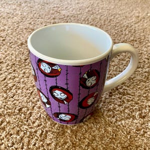 Puede incluir: Una taza de cer&aacute;mica blanca con un exterior morado que presenta un patr&oacute;n repetido de la cara de un personaje de la pel&iacute;cula "Pesadilla antes de Navidad". La taza tiene un asa blanca y un interior blanco.