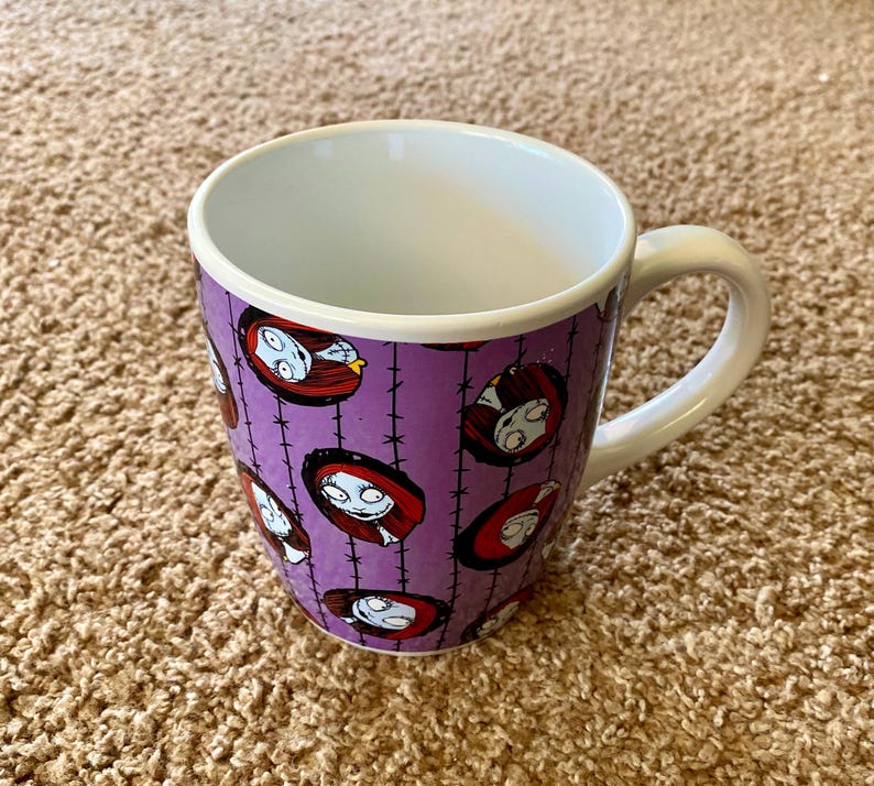 Puede incluir: Una taza de cer&aacute;mica blanca con un exterior morado que presenta un patr&oacute;n repetido de la cara de un personaje de la pel&iacute;cula "Pesadilla antes de Navidad". La taza tiene un asa blanca y un interior blanco.