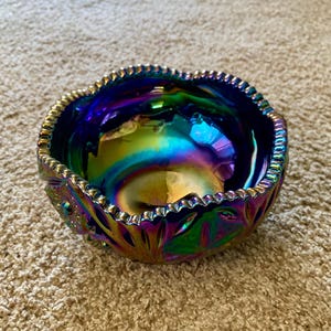 Puede incluir: Un cuenco de vidrio iridiscente con borde ondulado. El cuenco tiene un color base azul oscuro con un brillo arcoíris de verde, morado y dorado. El cuenco tiene un patrón floral decorativo.