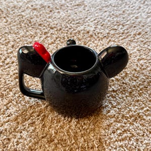 May include: Black ceramic mug shaped like a character's head, featuring large ears and a red bow. The mug has a handle and a glossy finish, perfect for hot beverages.